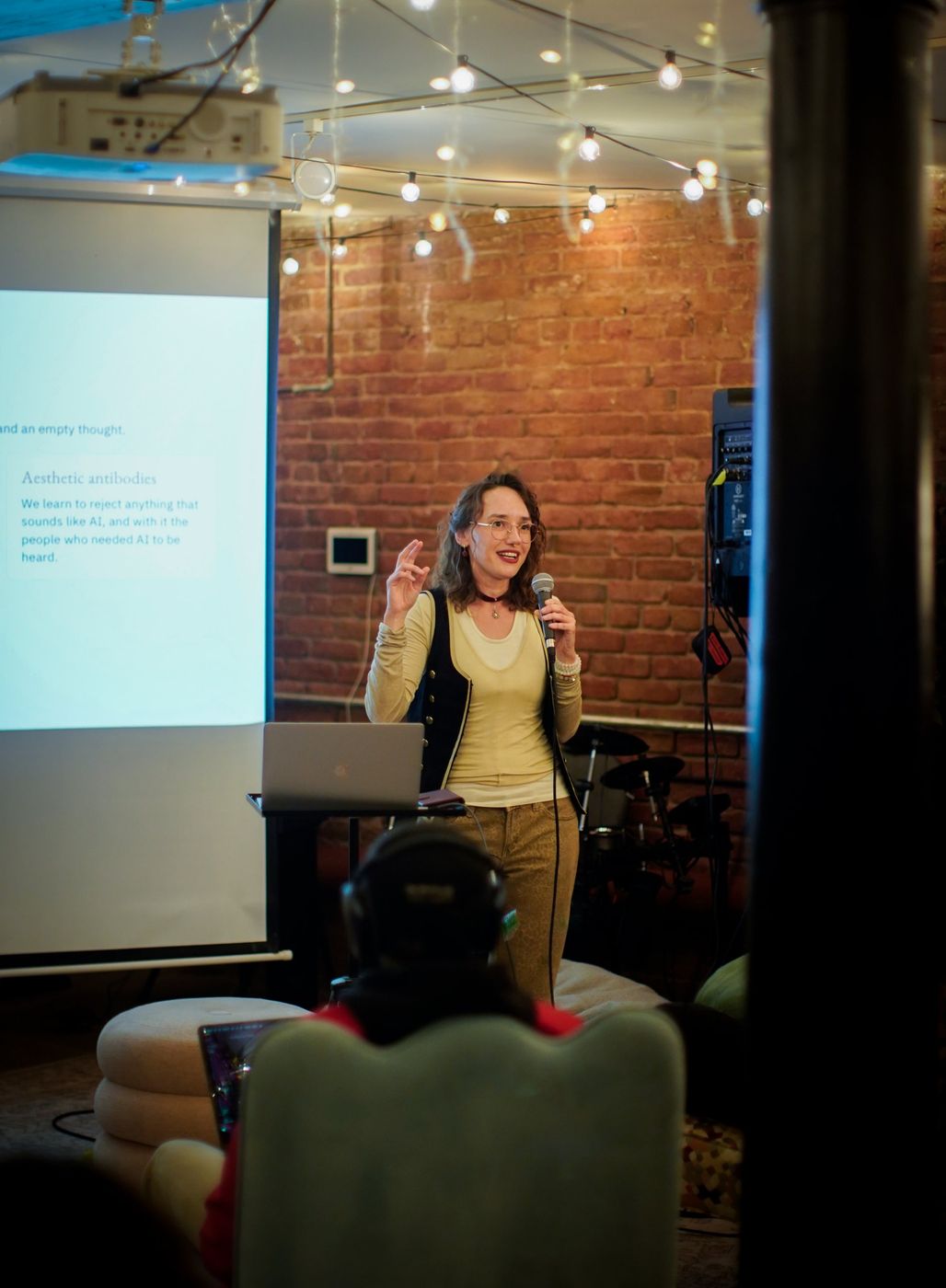 Anna speaking into a microphone beside a projected slide titled “Aesthetic antibodies”, with a laptop on a stand in front of a brick wall