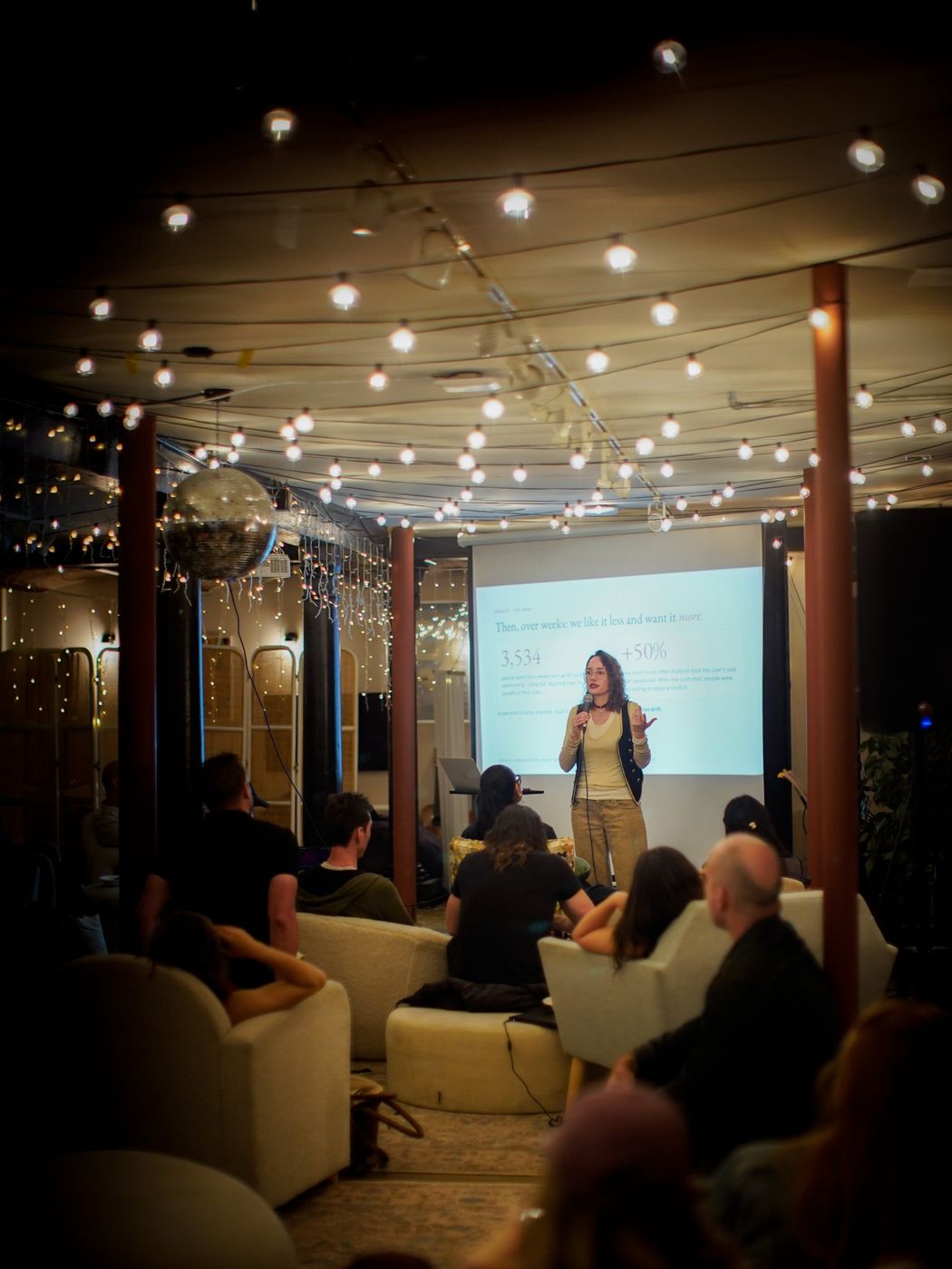 A wide view of The Commons: a speaker with a microphone beside a projected slide, the audience in armchairs under string lights and a disco ball