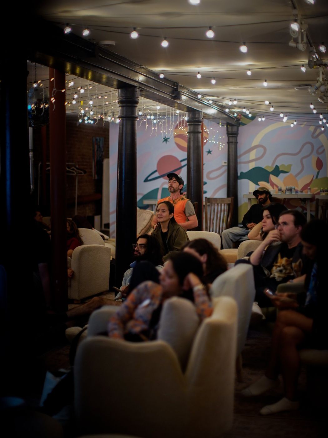 The audience seated in armchairs and on cushions under string lights, facing the screen, with a painted mural of planets behind them