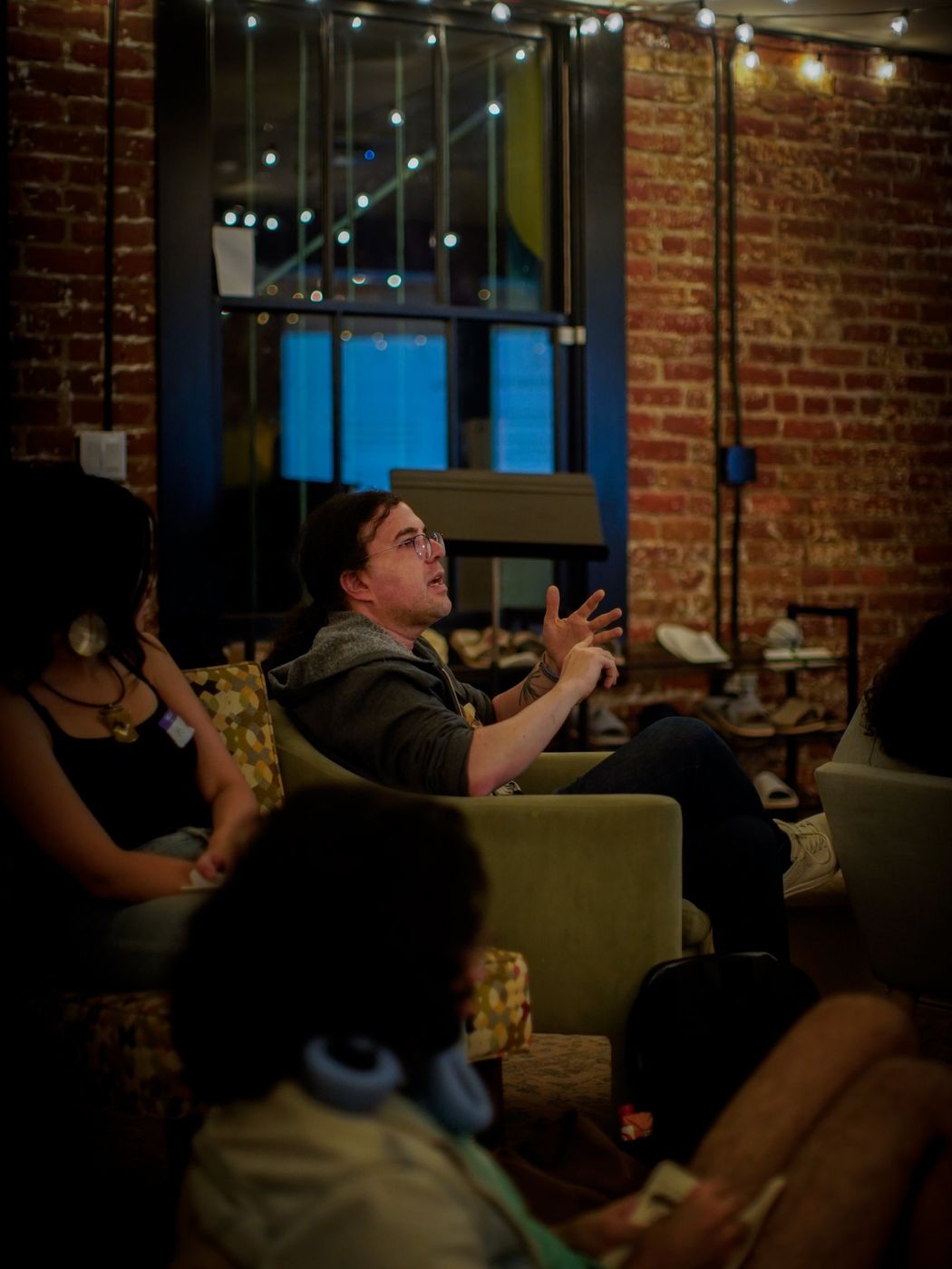 A man in a hooded sweatshirt gesturing as he speaks, seated in a green armchair among other attendees
