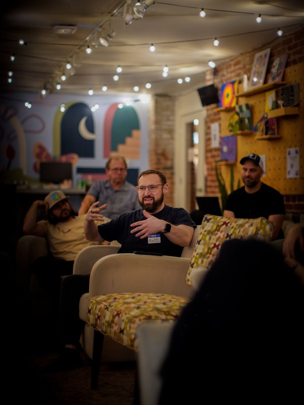 A bearded man in glasses gesturing as he speaks, with other attendees seated behind him