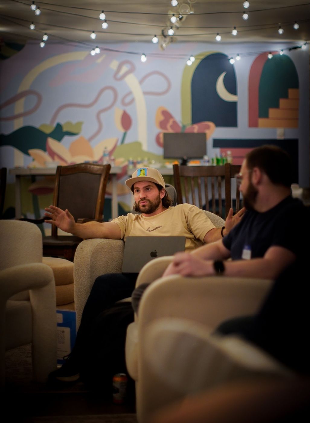 Two men talking in armchairs, one with a laptop on his lap, in front of a painted mural