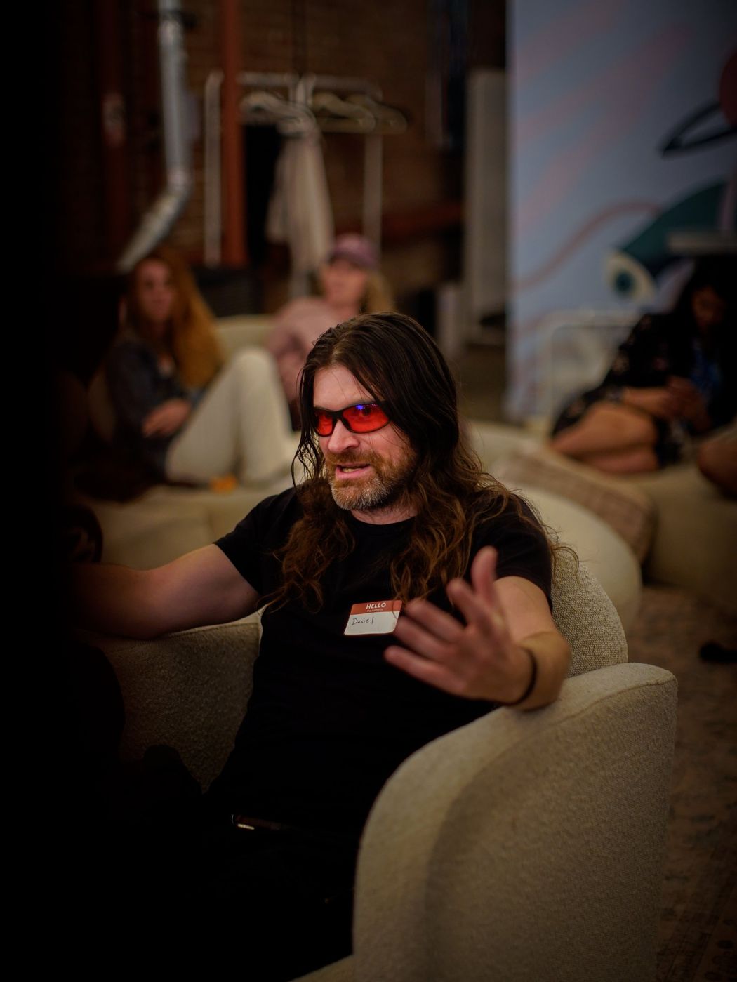A long-haired man in orange-tinted glasses gesturing as he talks, seated in an armchair