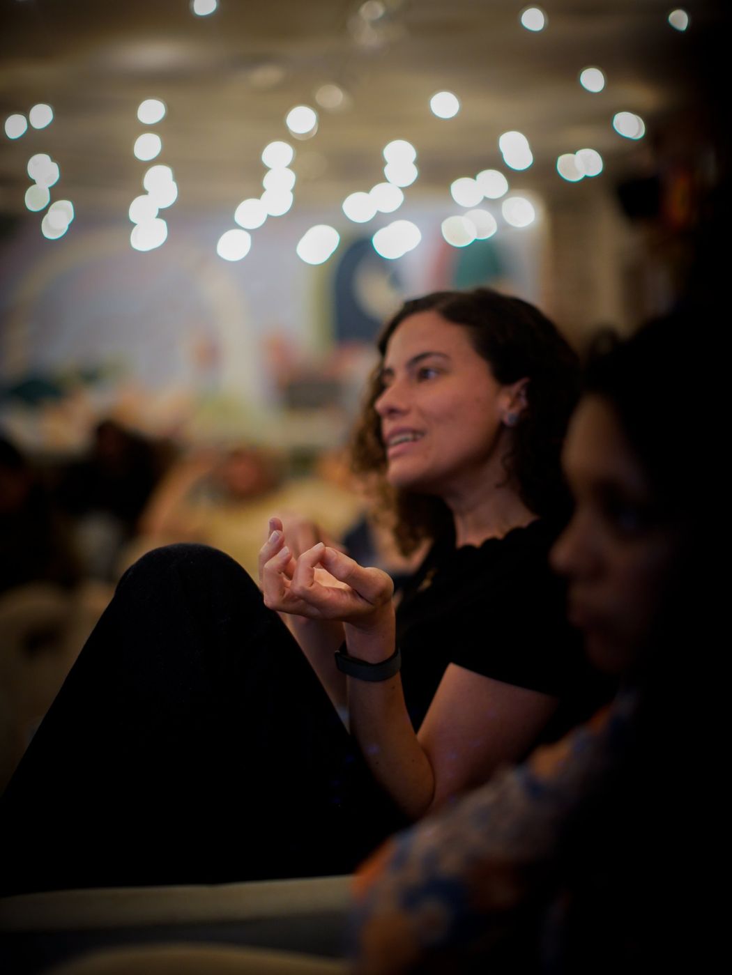 A woman with curly hair speaking, with string lights softly blurred behind her