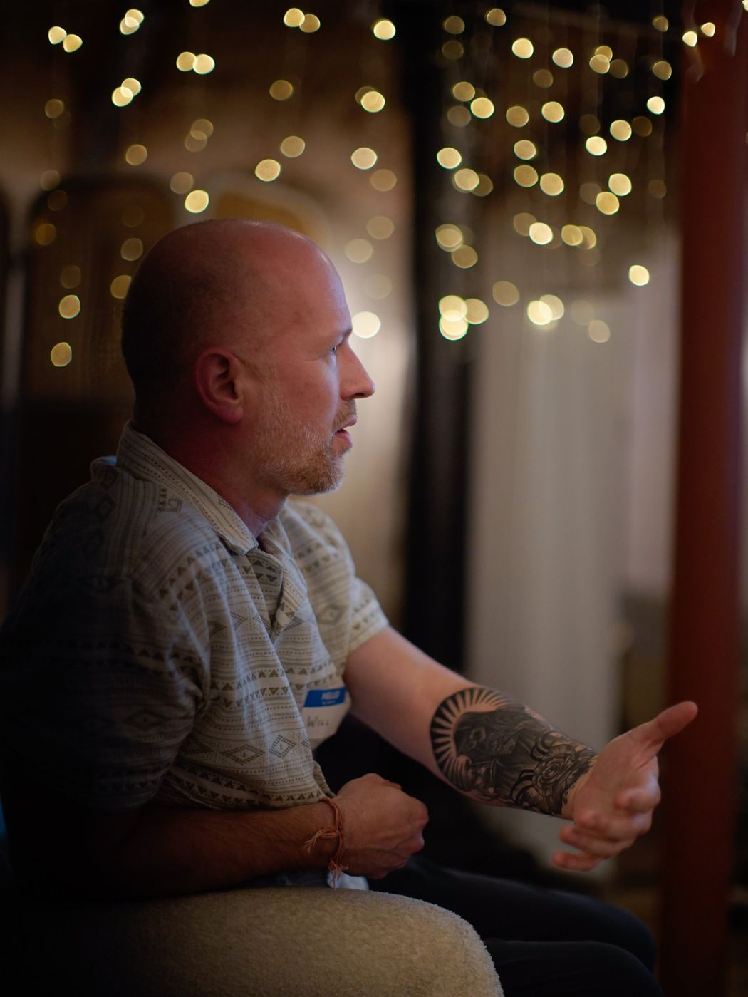A man in profile gesturing with a tattooed forearm as he speaks, with fairy lights behind him