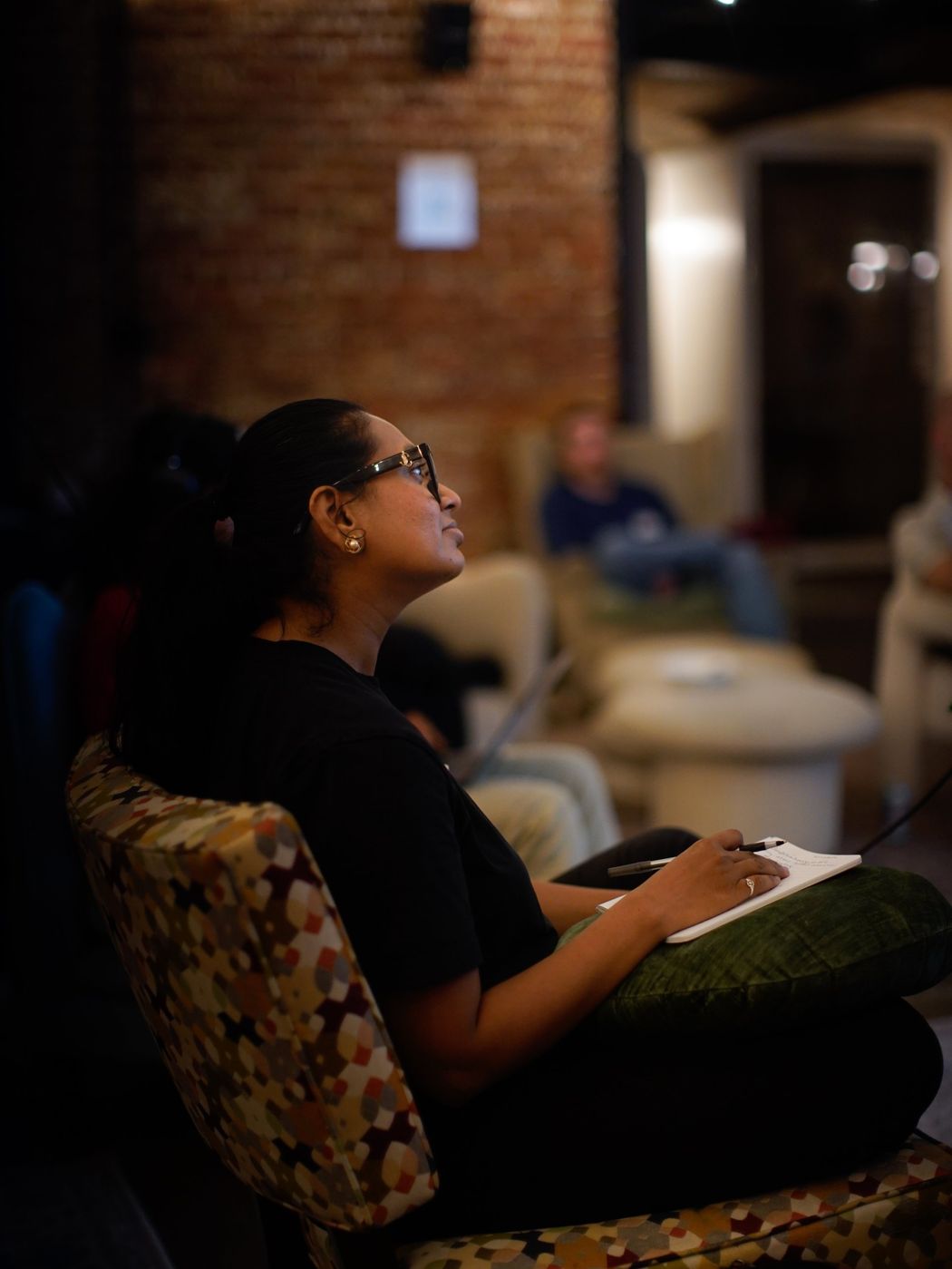 A woman in glasses seated in a patterned armchair, looking up at the screen with a notebook on her lap