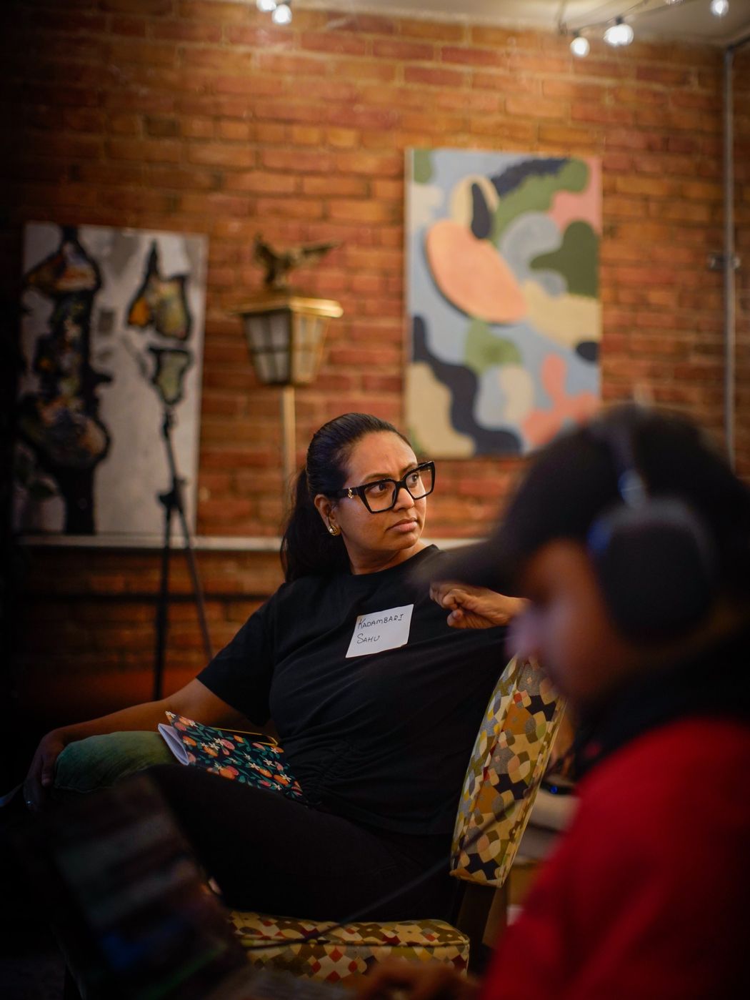 A woman in black-framed glasses seated in an armchair, looking across the room, with abstract paintings on the brick wall behind her