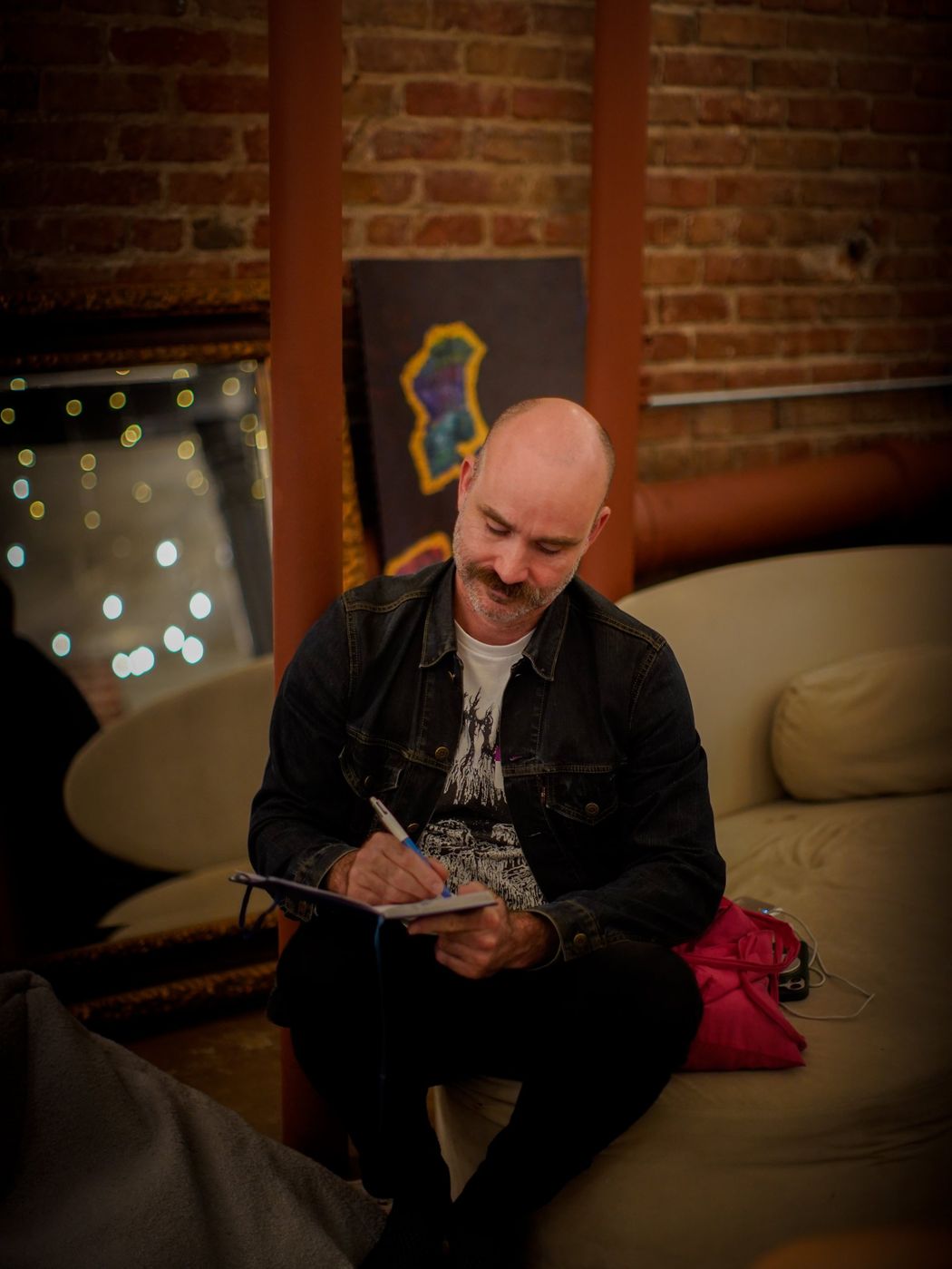 A man in a denim jacket writing in a notebook, seated on a couch against a brick wall