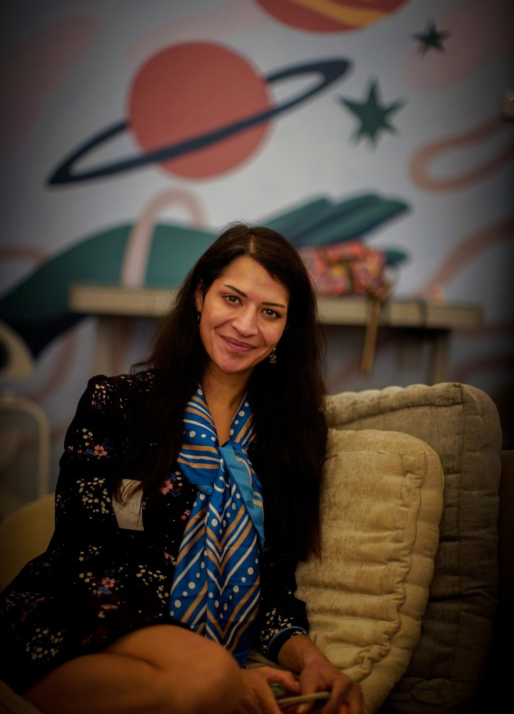A woman with long dark hair and a blue scarf smiling at the camera, seated in front of a planet mural