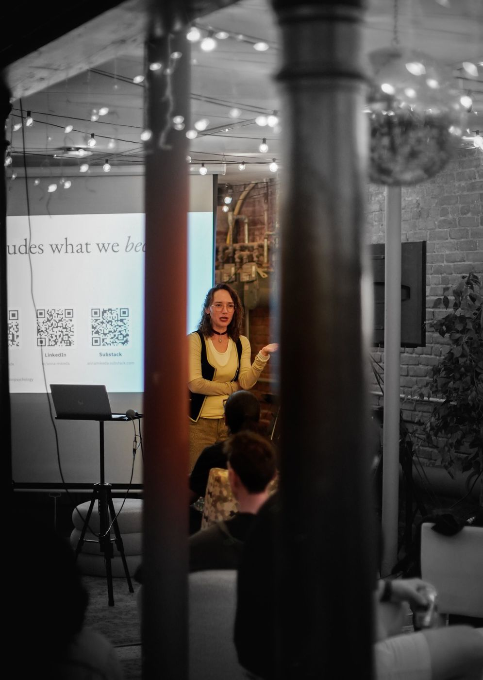 Anna speaking in front of a slide with QR codes, seen between two pillars; the room is in black and white and the speaker in colour